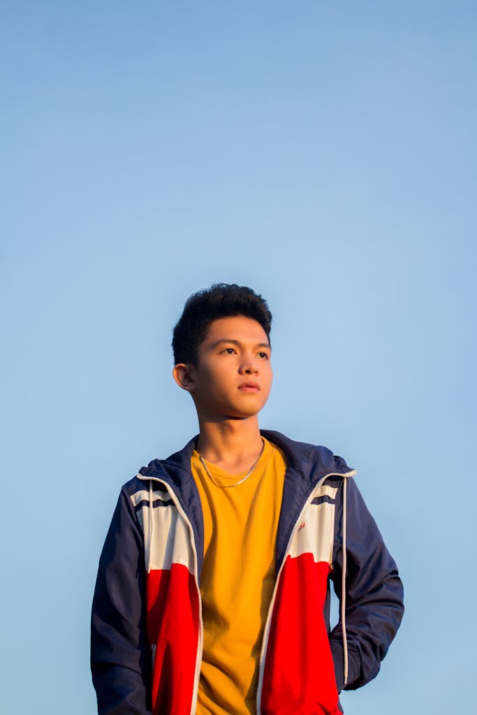 Portrait of a teenage boy in a colorful jacket looking away with a clear sky background.
